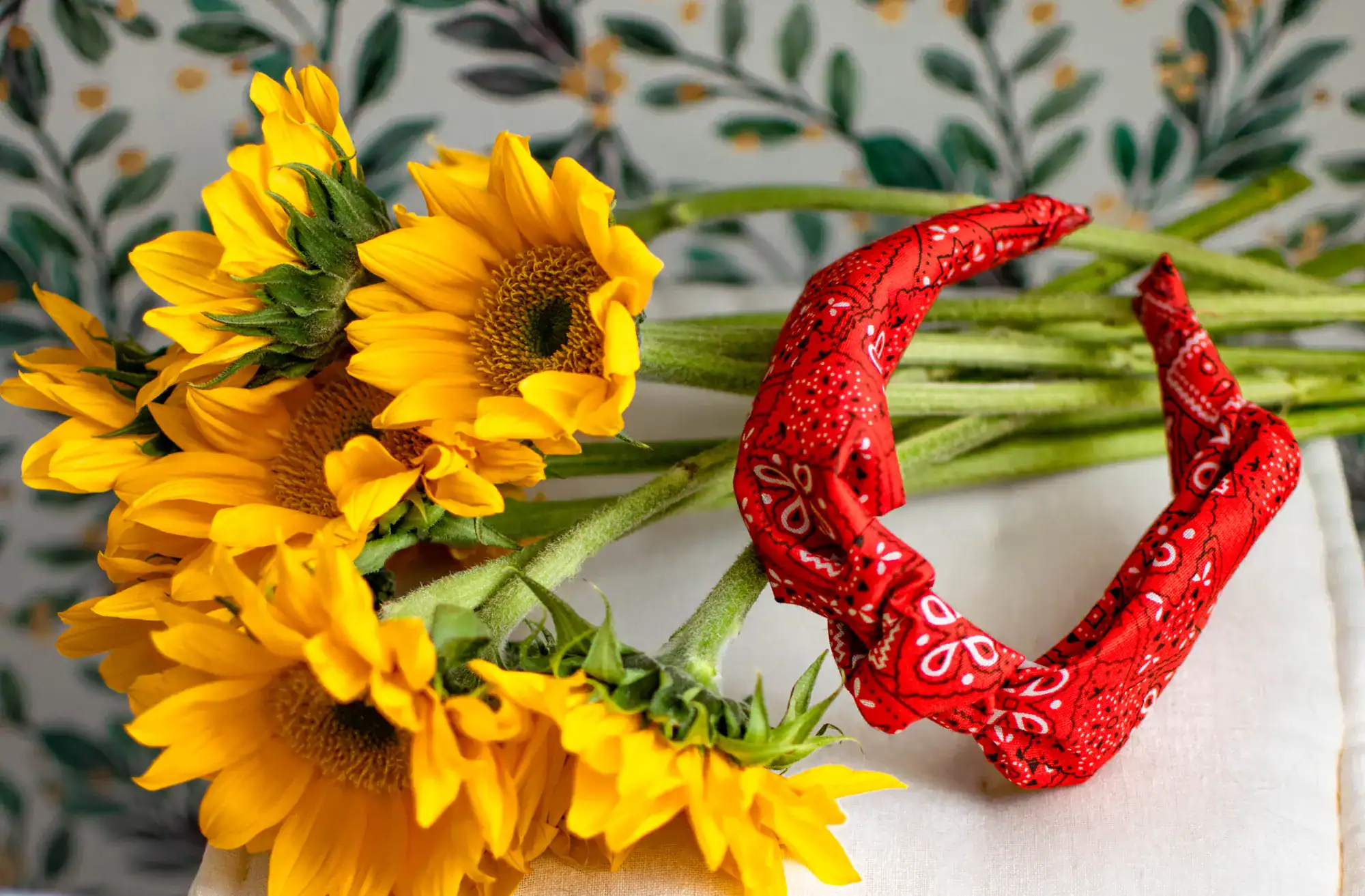 Red bandana print turban headband beside a bouquet of bright yellow sunflowers on a white surface with a leafy patterned background.