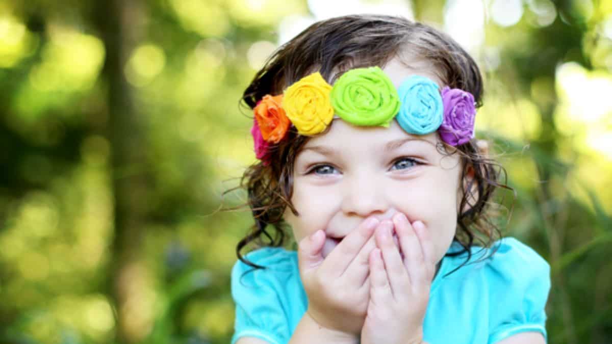 Young girl wearing a rainbow fabric flower headband with colorful rolled flowers, smiling outdoors in a green garden setting.