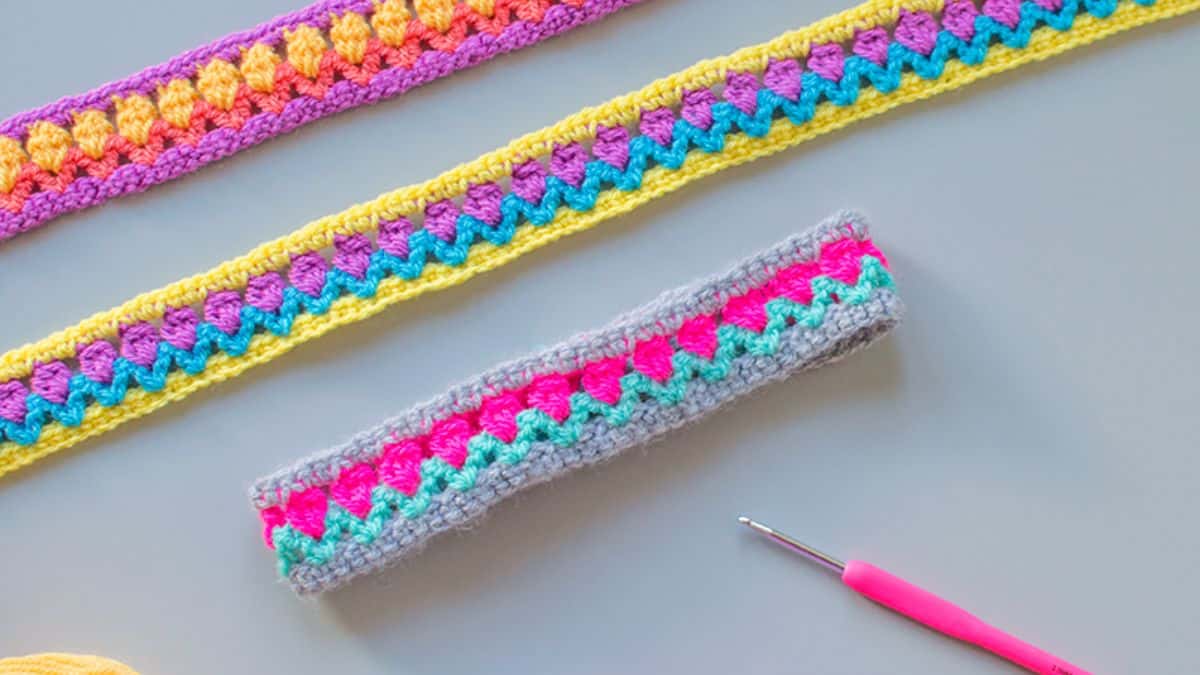 Colorful crochet headbands with zigzag and tulip stitch patterns displayed on a gray surface beside a pink crochet hook.