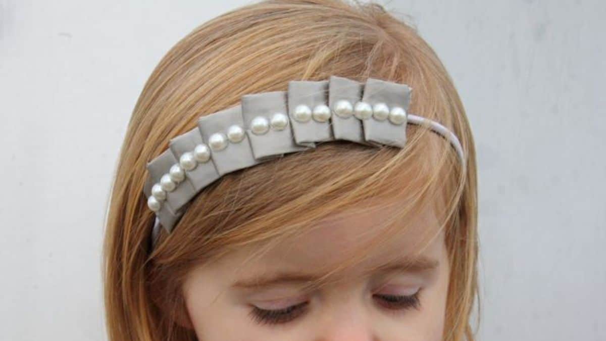 Young girl wearing a gray pleated headband decorated with pearl accents and small fabric bows.