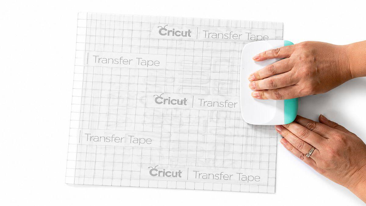 Hands using a Cricut scraper tool to smooth transfer tape over a vinyl design, helping the decal adhere evenly before application.