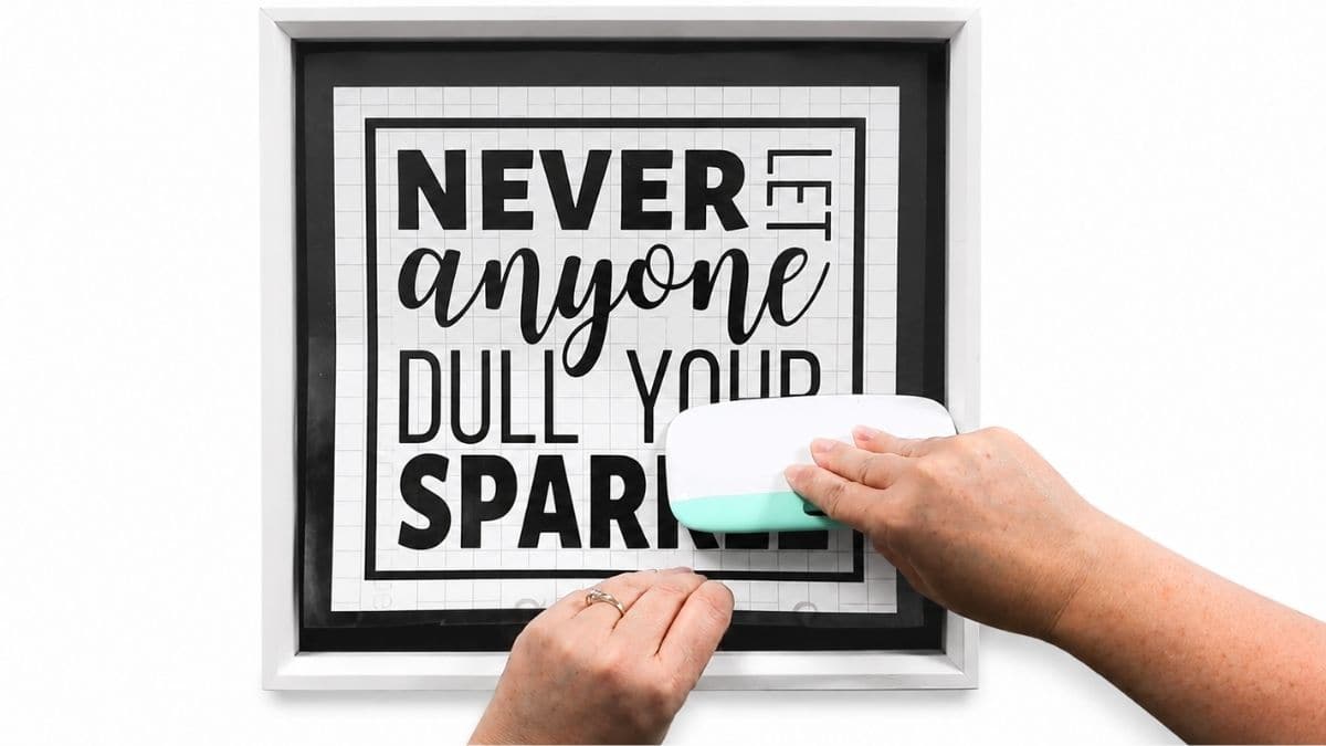 Hands using a Cricut scraper tool to smooth a vinyl quote decal onto a white shadow box frame during a DIY glitter sign project.
