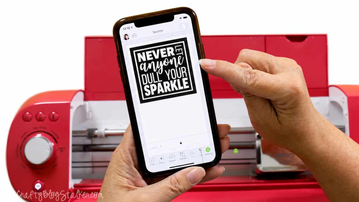 Hands holding a smartphone displaying a Cricut Design Space quote design in front of a red Cricut cutting machine.