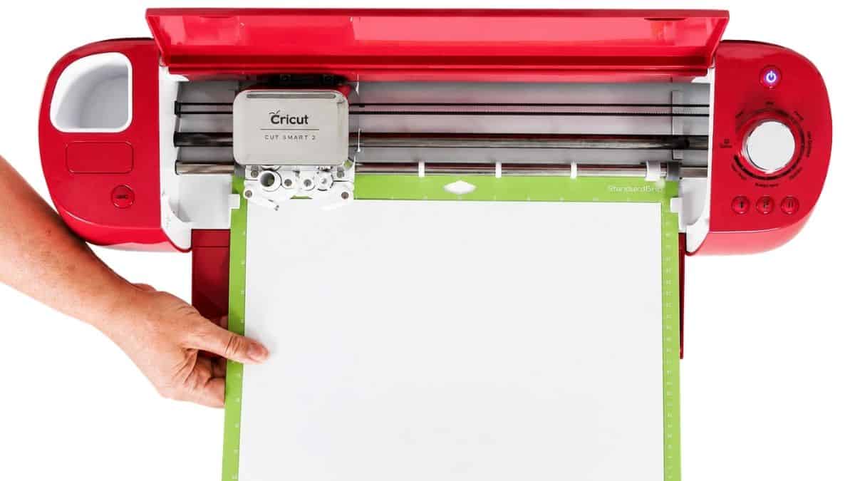 Hand loading a Cricut cutting mat with white cardstock into a red Cricut machine, ready to cut a vinyl or paper design.