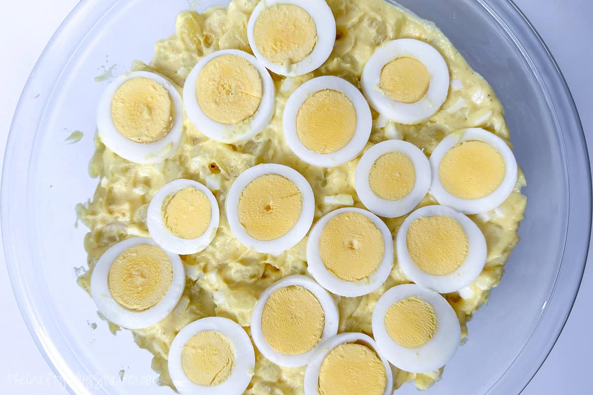 Close-up of creamy potato salad in a glass bowl, topped with evenly sliced hard-boiled eggs arranged in a circular pattern.