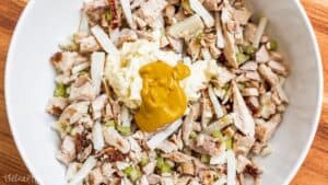 Rotisserie chicken, jicama, and celery topped with mayonnaise and mustard in a white mixing bowl before being mixed into chicken salad.