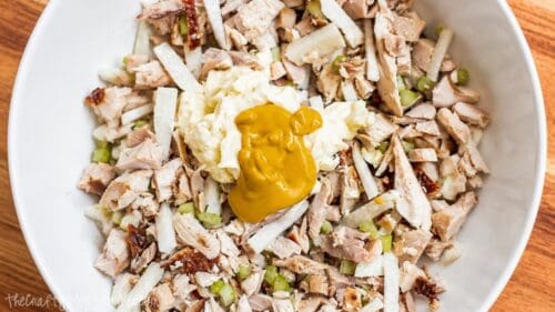 Rotisserie chicken, jicama, and celery topped with mayonnaise and mustard in a white mixing bowl before being mixed into chicken salad.