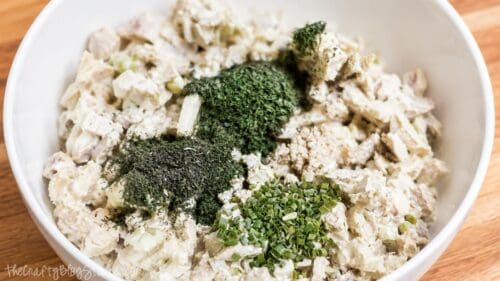 Rotisserie chicken salad ingredients in a white bowl topped with fresh parsley, dill, chives, and minced garlic before mixing.