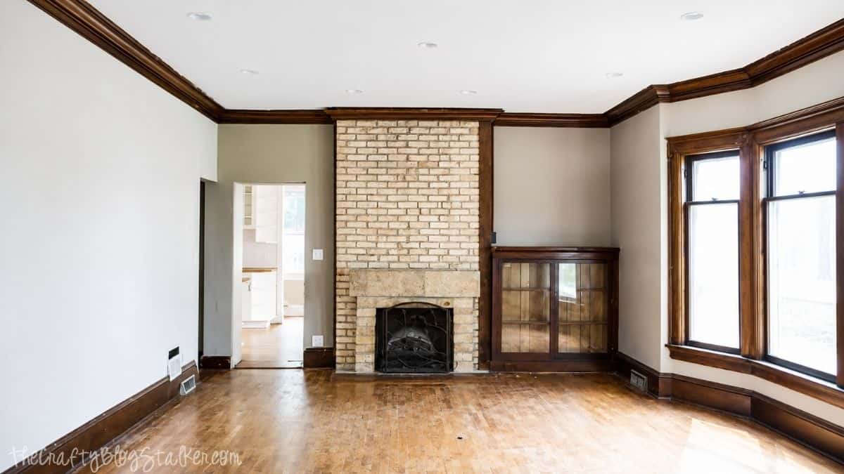 Empty living room with hardwood floors, a painted brick fireplace, built-in cabinet, and large wood-trimmed windows before renovation.