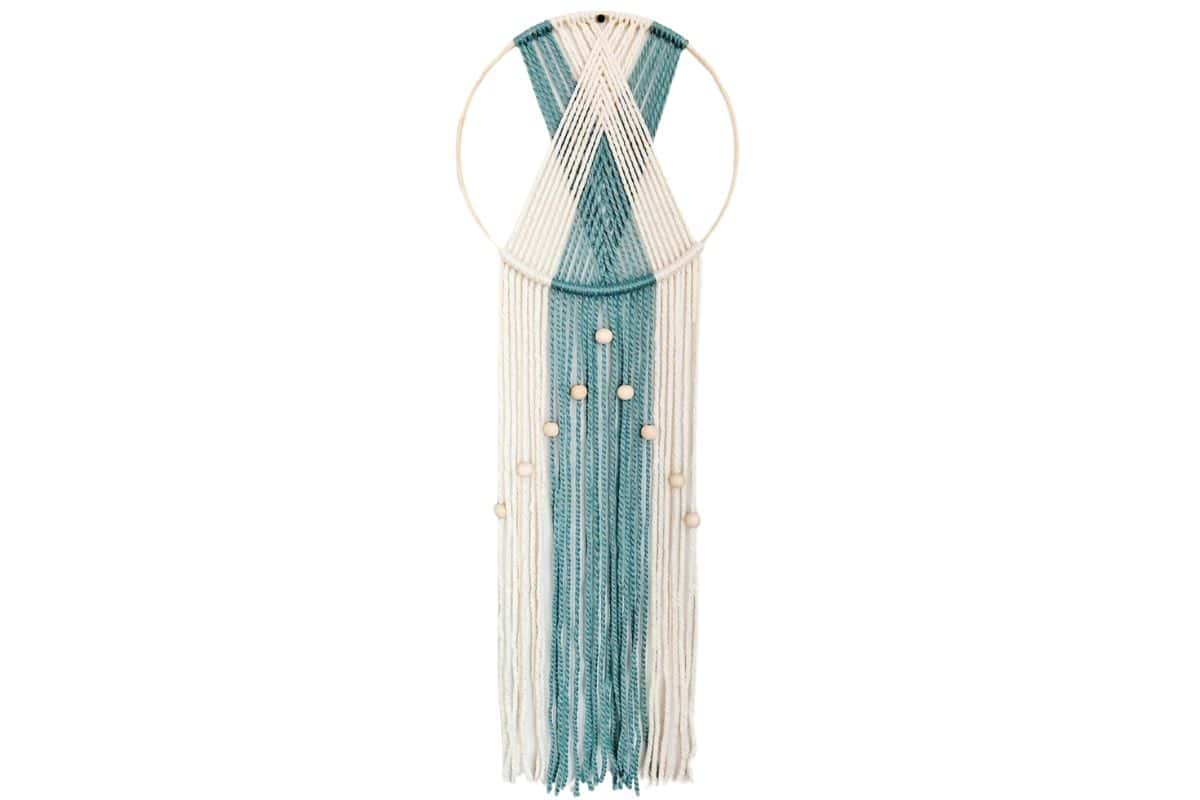 Modern yarn wall hanging with cream and teal strands, arranged in a chevron pattern on a circular frame and decorated with wooden beads.