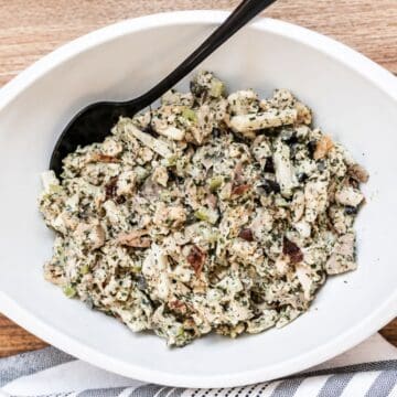 Creamy rotisserie chicken salad with chopped chicken, celery, grapes, almonds, herbs, and dressing served in a white bowl with a black serving spoon.
