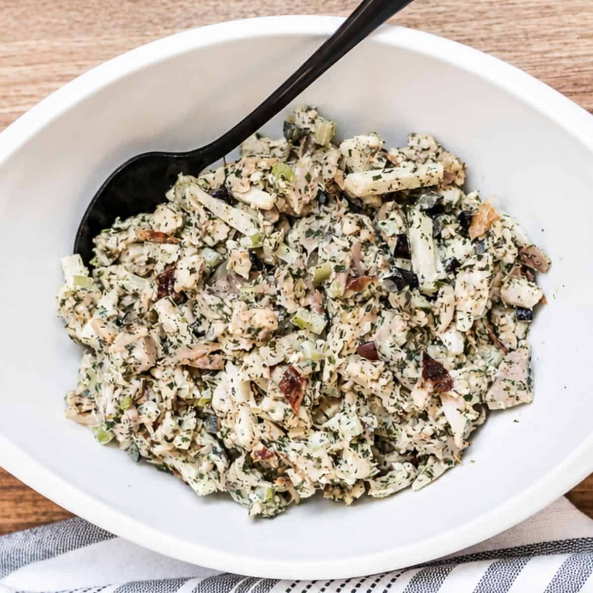 Creamy rotisserie chicken salad with chopped chicken, celery, grapes, almonds, herbs, and dressing served in a white bowl with a black serving spoon.