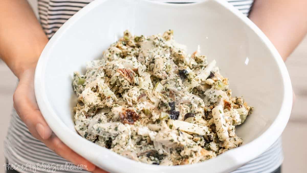 Person holding a large white bowl filled with creamy rotisserie chicken salad made with chicken, jicama, celery, herbs, and grapes.