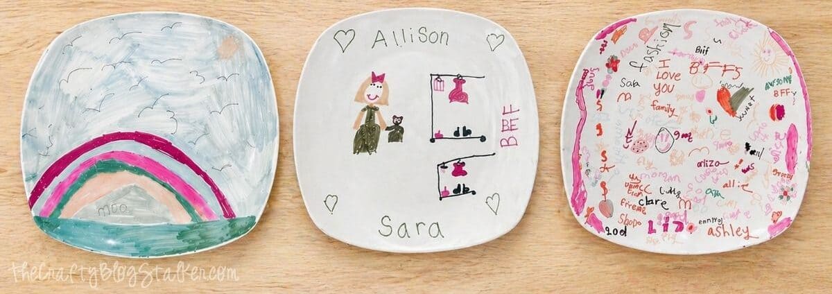 Three white ceramic plates decorated with colorful Sharpie drawings, including a rainbow, names, and doodles, displayed on a wooden table.