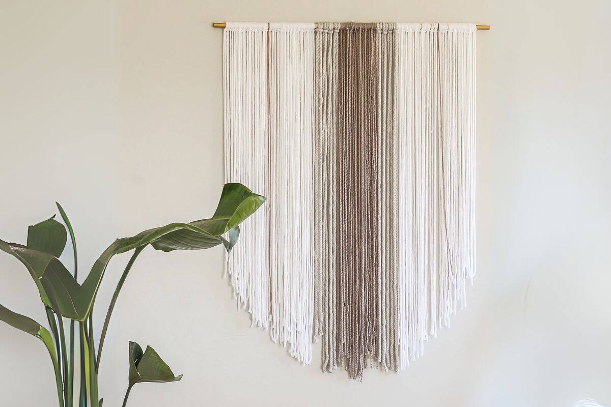 Neutral-toned yarn wall hanging in cream and taupe colors, draped in a chevron shape from a gold rod, displayed above a large leafy green houseplant.