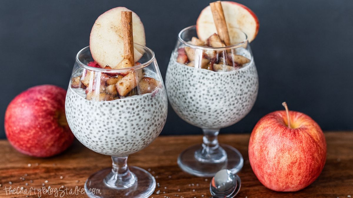 Two glasses of chia pudding topped with cinnamon apples, apple slices, and cinnamon sticks on a wooden surface with fresh apples beside them.