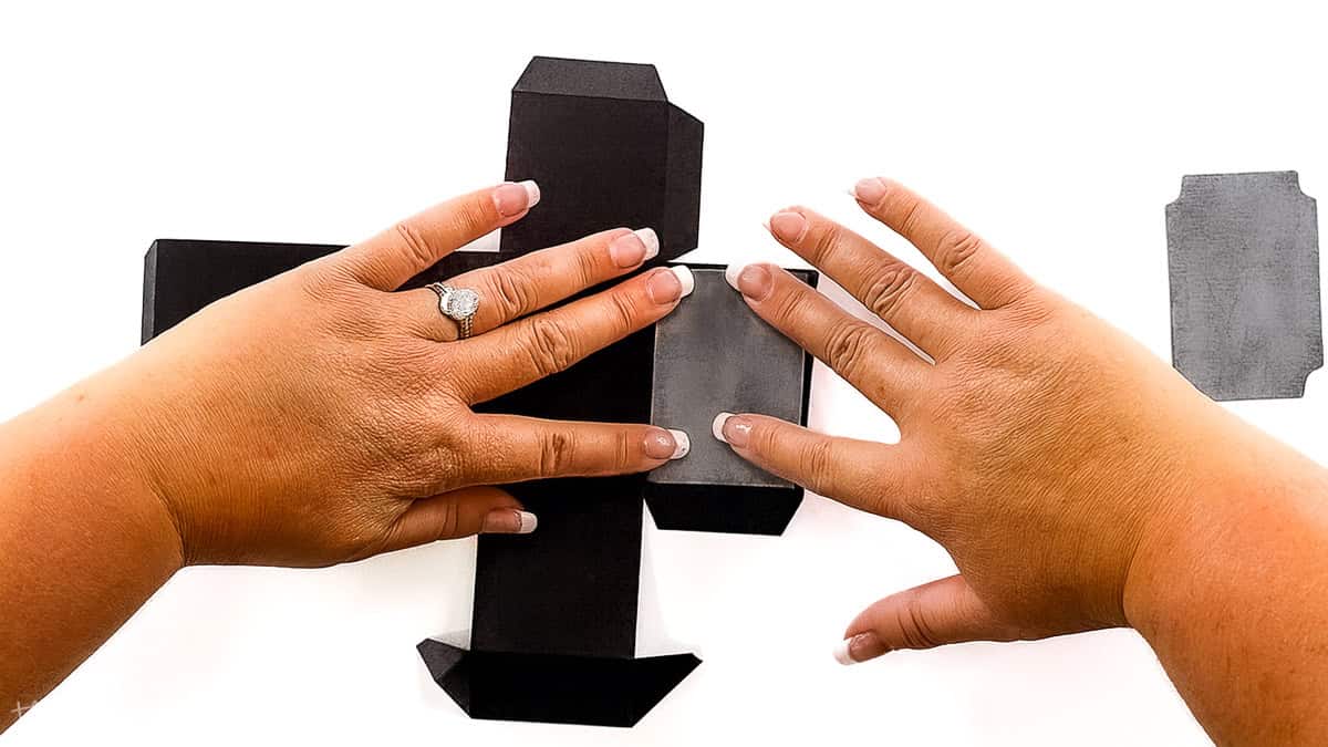 Hands assembling a black paper box and pressing gray panels onto the sides to create a Halloween gravestone candy holder craft.