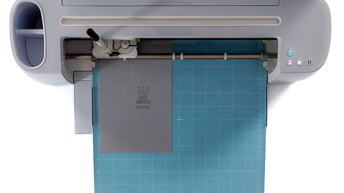Cricut cutting machine engraving a gravestone design on black cardstock with the text 