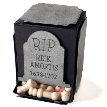 DIY black paper gravestone candy dispenser labeled “RIP Rick Amortis 1678–1702,” filled with bone-shaped candies at the base.