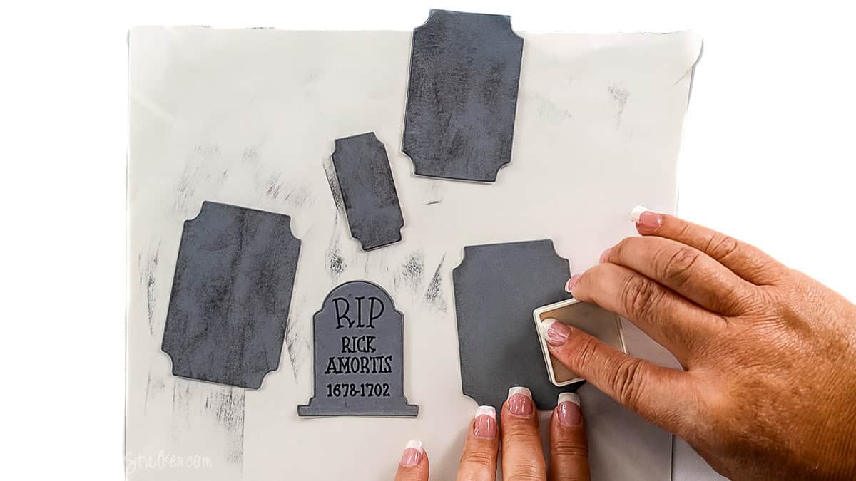 Hands using an ink pad to distress gray paper gravestone pieces labeled 