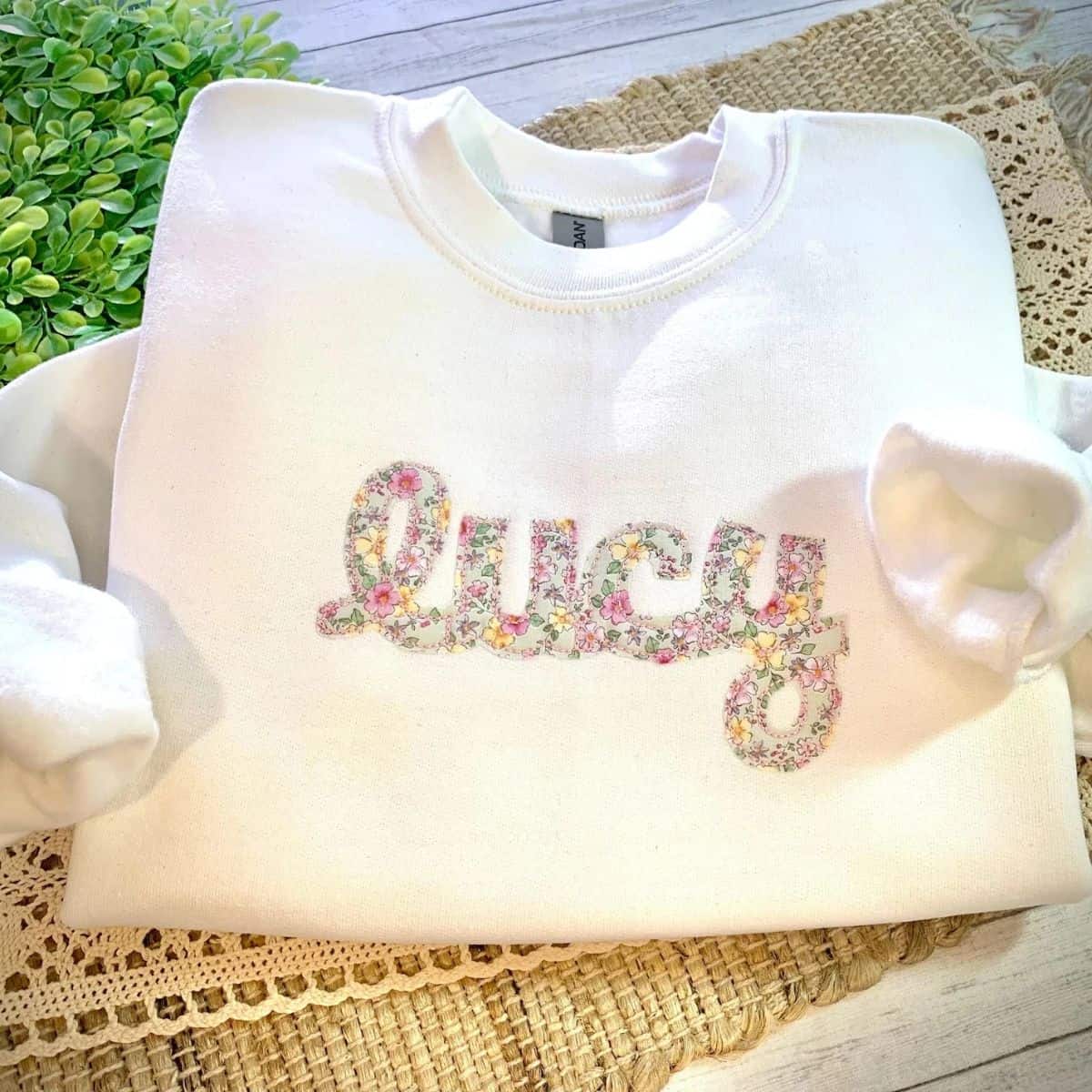 White crewneck sweatshirt personalized with the name Lucy in pastel floral lettering, displayed on a woven mat with lace trim.