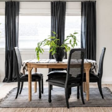 Modern dining room with a wood table, black mesh-back chairs, layered rugs, black curtains, and a leafy plant centerpiece.