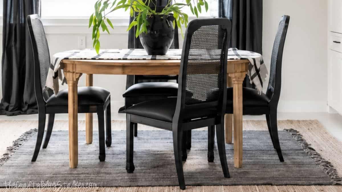 Wood dining table with black dining chairs, layered area rugs, and a black vase with greenery in a bright modern dining room.