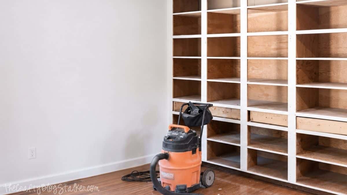 Built-in cabinet frames with doors removed during a dining room renovation, with a shop vacuum on hardwood floors beside unfinished shelving.