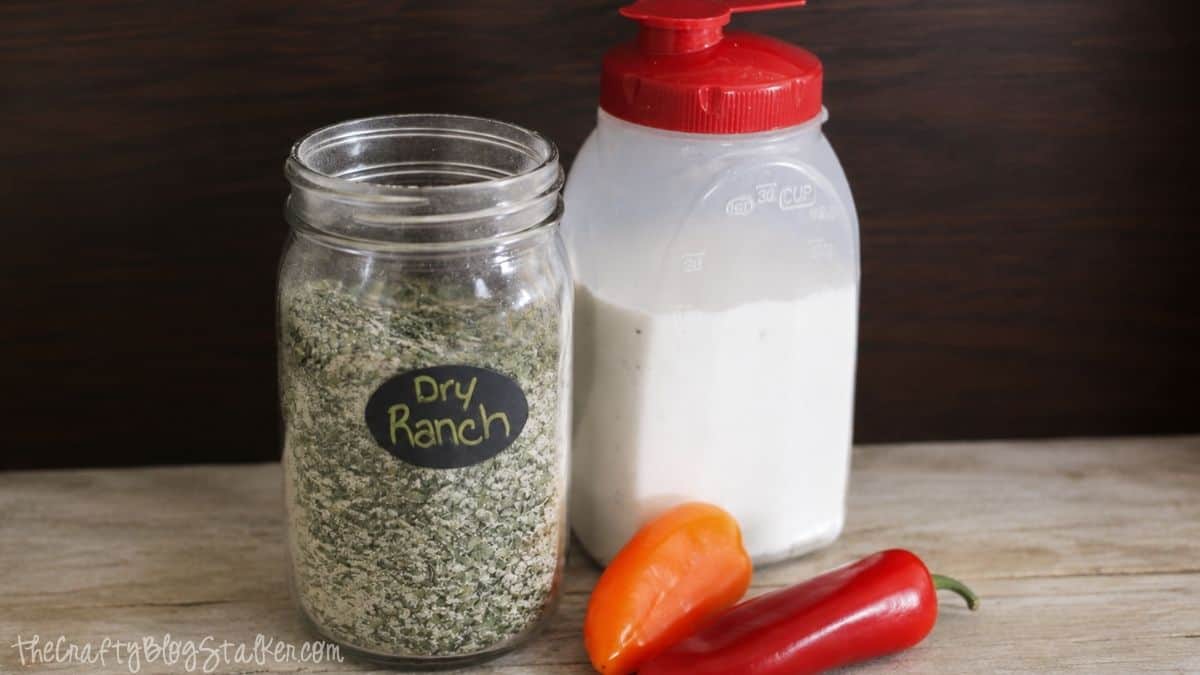 Jar of homemade dry ranch seasoning mix beside a container of buttermilk with red and orange mini peppers on a wooden tabletop.