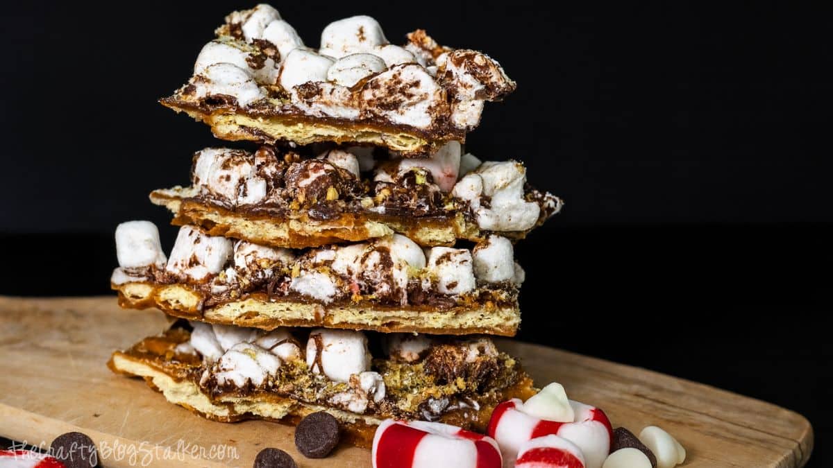 Stack of peppermint s'mores toffee topped with mini marshmallows, chocolate, and crushed candy pieces on a wooden board against a dark background.