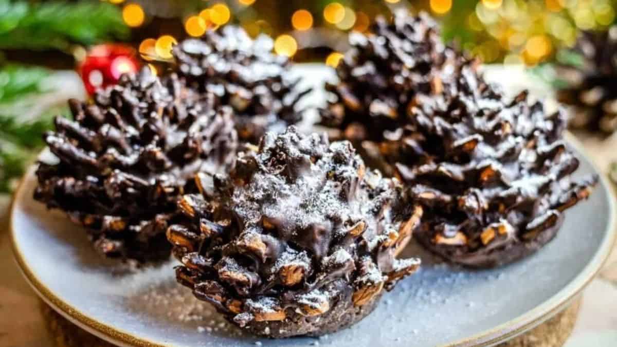 Chocolate pinecone-shaped desserts made from cereal and cocoa, dusted with powdered sugar and arranged on a plate with festive holiday lights in the background.