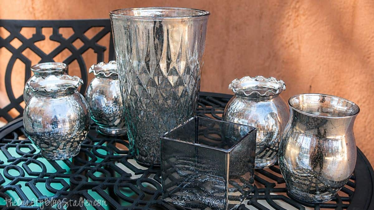 Assorted glass vases and candle holders with a mercury glass finish displayed on a black metal table outdoors.