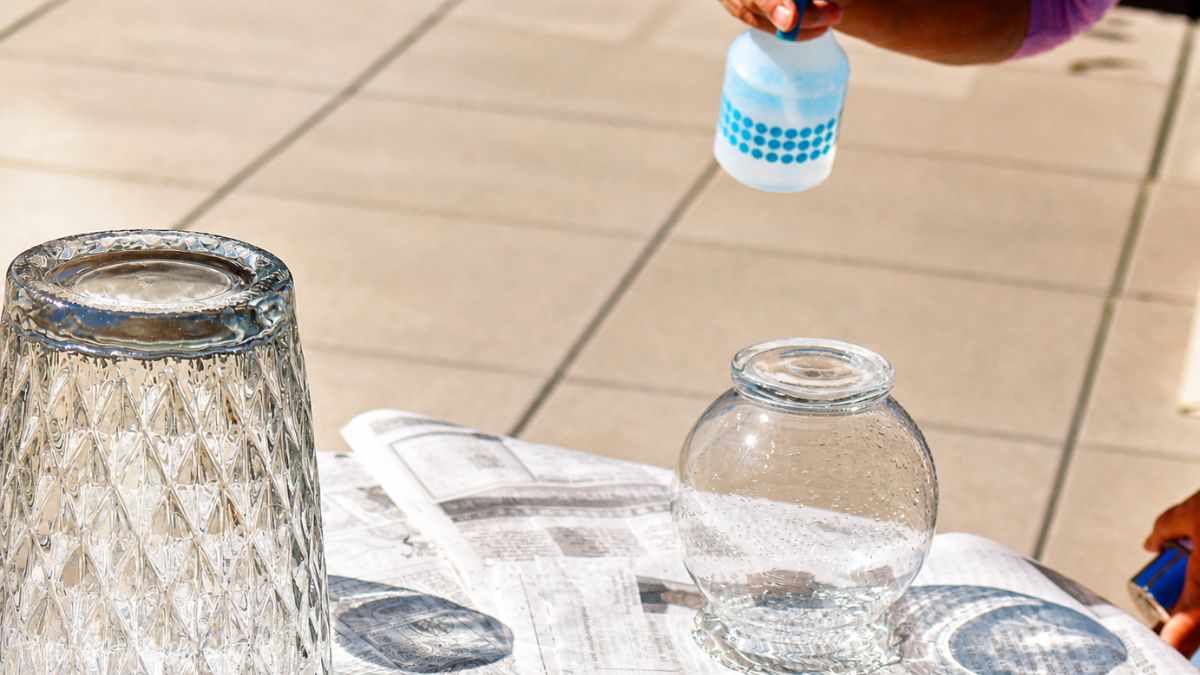 Hand spraying water onto clear glass jars set on newspaper, preparing the surface for a DIY mercury glass finish.