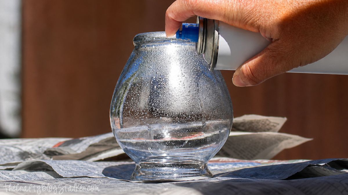 Hand spraying silver paint onto a water-speckled glass jar on newspaper to create a DIY mercury glass effect.