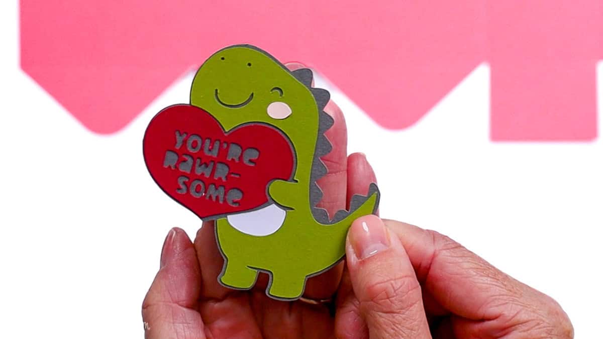 Hands holding an assembled paper dinosaur holding a heart with the phrase