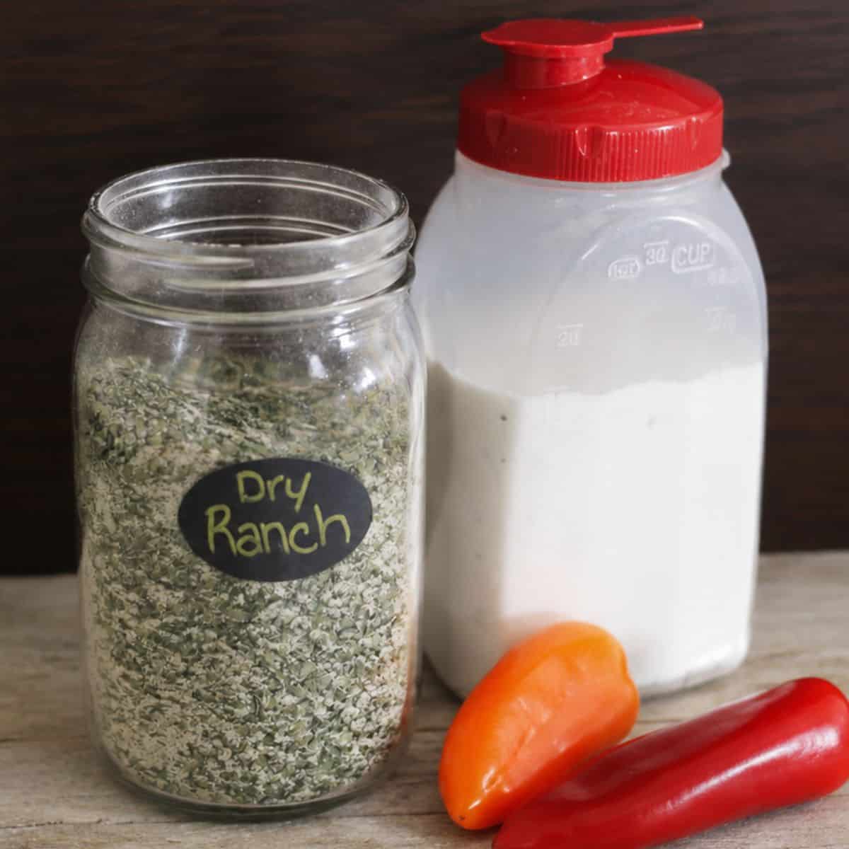 Jar of homemade dry ranch seasoning mix beside a container of buttermilk with red and orange mini peppers on a wooden tabletop.