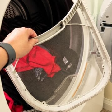 Hand removing a large lint screen from an open clothes dryer with laundry visible inside.