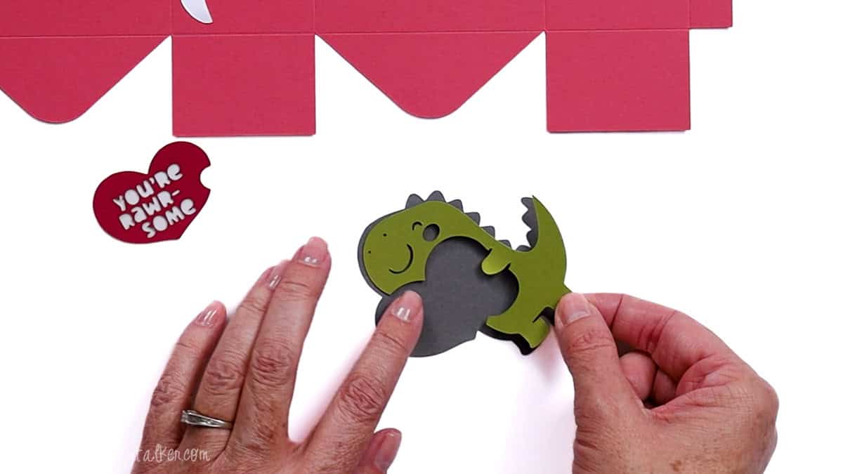 Hands assembling layered paper dinosaur pieces with a heart cutout, shown below flat cardstock panels for a Valentine treat bag project.