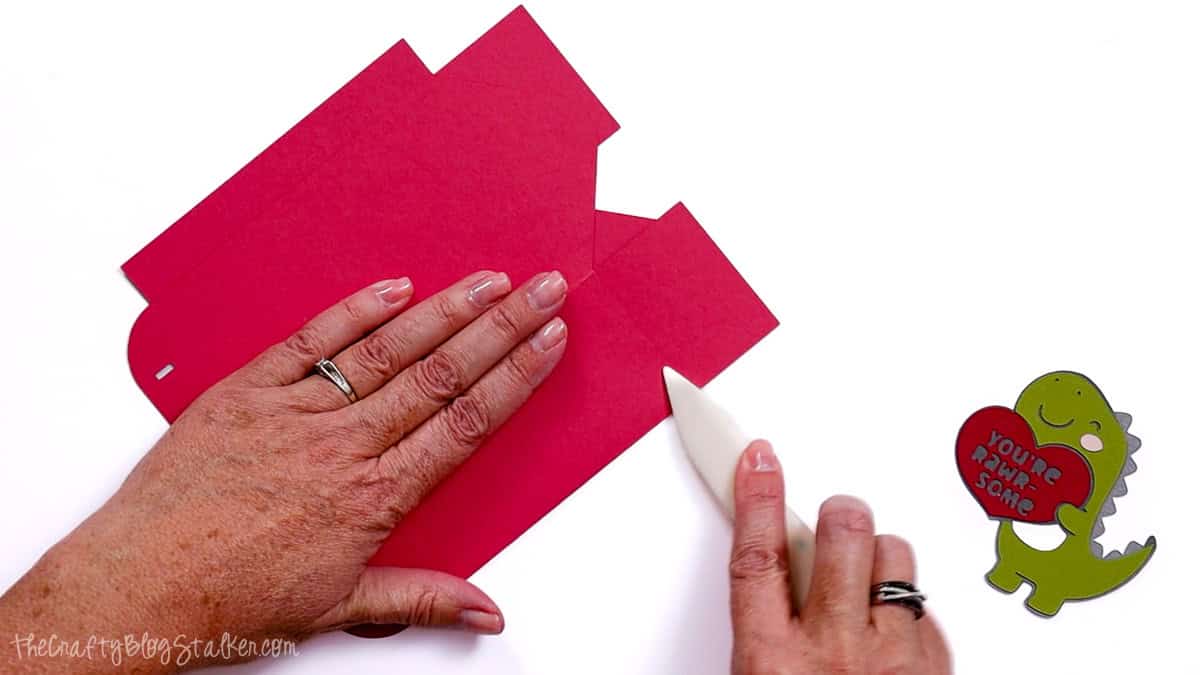 Hands using a bone folder to score and fold a cardstock treat bag template, with a finished dinosaur Valentine embellishment beside it.