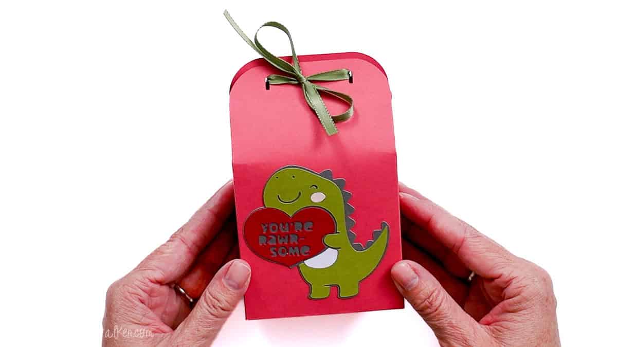 Finished dinosaur Valentine treat bag made from cardstock, decorated with a layered dinosaur holding a heart and tied closed with a ribbon.