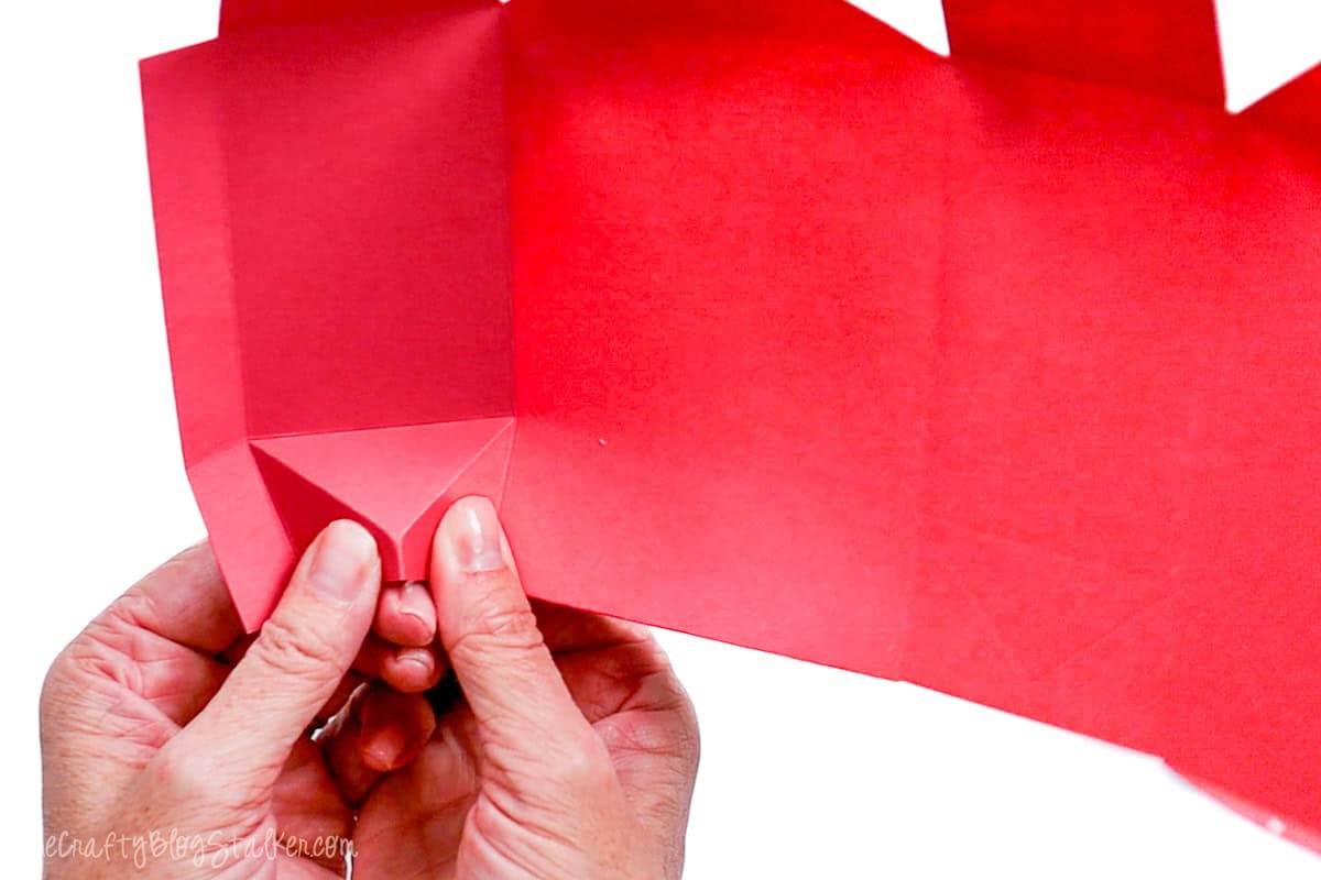 Hands pinching and folding the top corners of a cardstock treat bag to form the base during assembly.
