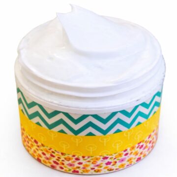 Small jar of white body lotion with decorative patterned labels around the container and a smooth swirl of lotion on top.