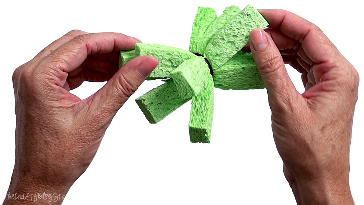 Hands fluffing and arranging the sponge strips on a finished green sponge water bomb to create its round shape.