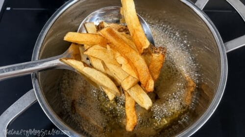 Slotted spoon lifting golden homemade French fries from hot oil in a large stainless steel pot after frying.