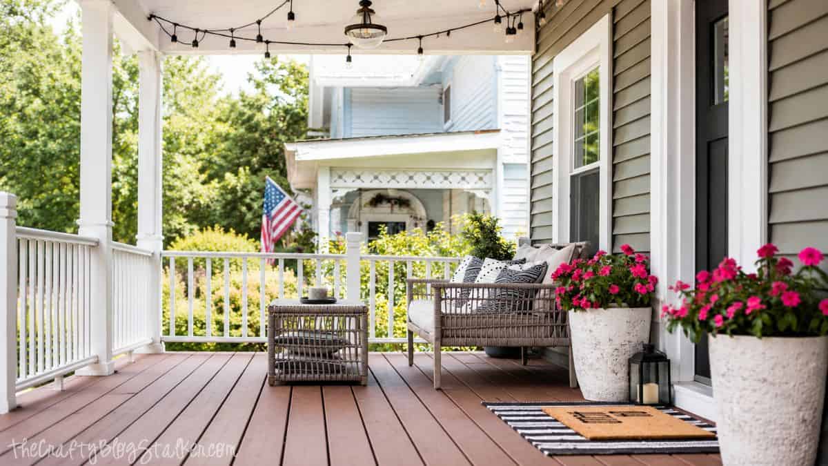 Covered front porch with wicker seating, string lights, potted pink flowers, and a striped welcome mat outside a gray house.