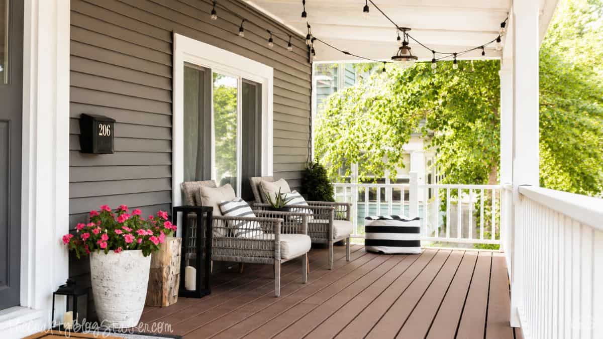 Front porch with wicker chairs, string lights, potted flowers, lanterns, and striped pillows outside a gray house on a sunny day.