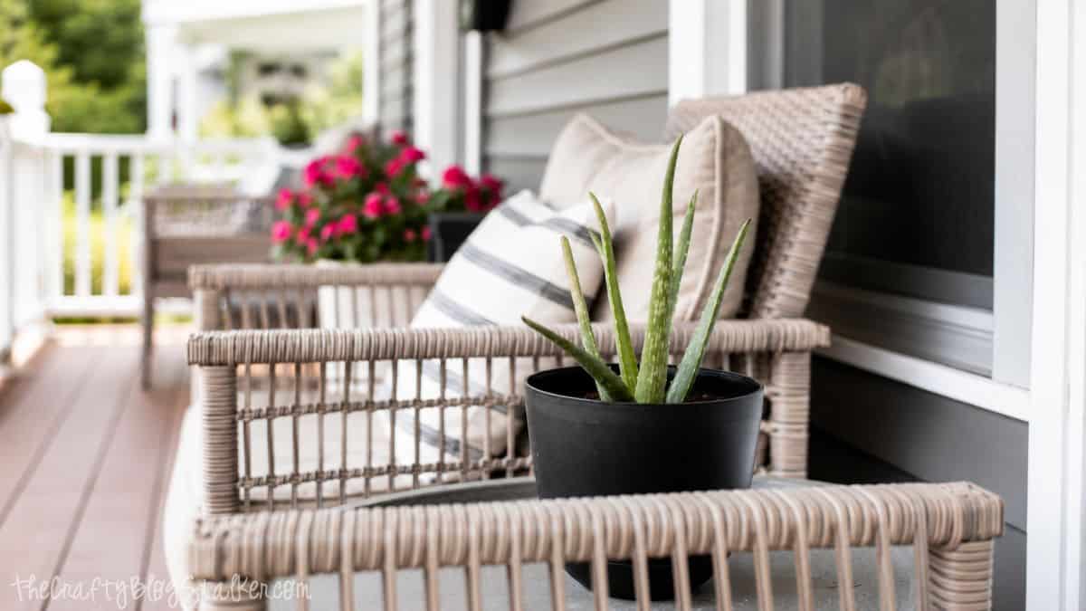 Close-up of wicker porch chairs with striped pillows and a small aloe plant on a covered front porch with flowers in the background.
