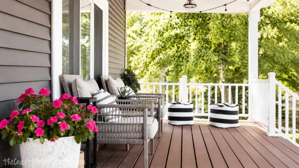Front porch with wicker chairs, striped pillows, potted pink flowers, and black-and-white poufs overlooking a green yard.