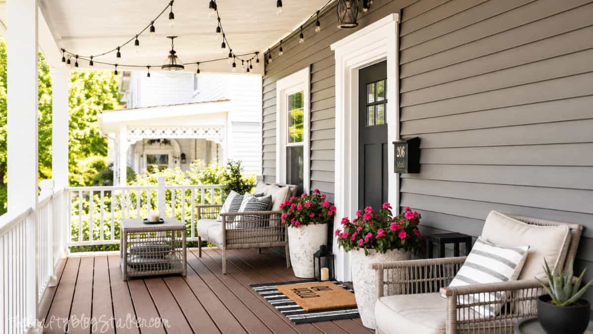 Wide covered front porch with wicker seating, string lights, pink flower planters, and striped pillows outside a gray house.