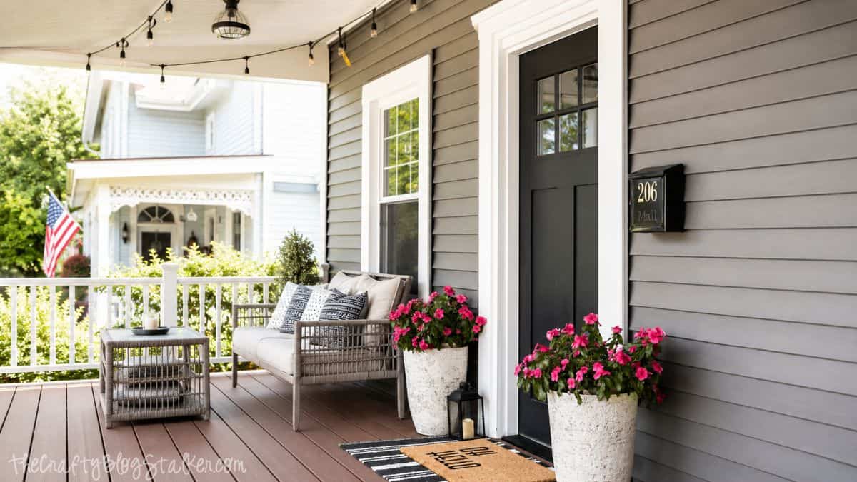 Covered front porch with wicker seating, string lights, pink flower planters, and a striped welcome mat outside a gray house.